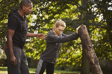 Young woman with personal trainer lifting tree trunk in park - CUF16379