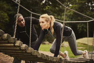 Personal trainer instructing young woman crawling on playground equipment in park - CUF16383