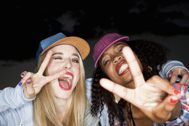 Young woman wearing baseball cap arm around friend looking at camera doing peace sign - CUF16517