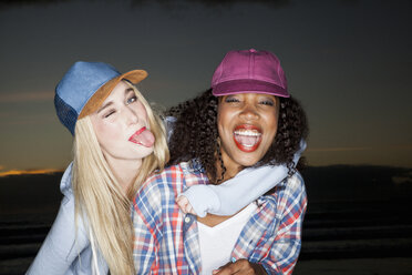 Young woman wearing baseball cap arm around friend looking at camera sticking out tongue - CUF16518