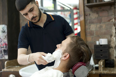 Junger Mann im Friseursalon trägt Rasierschaum auf das Gesicht eines Kunden auf - CUF16737
