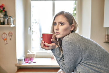 Junge Frau lehnt sich mit einer Tasse Kaffee an den Küchentisch - CUF16825