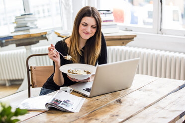 Junge Frau in einer Stadtwohnung frühstückt Müsli und liest am Laptop - CUF16844