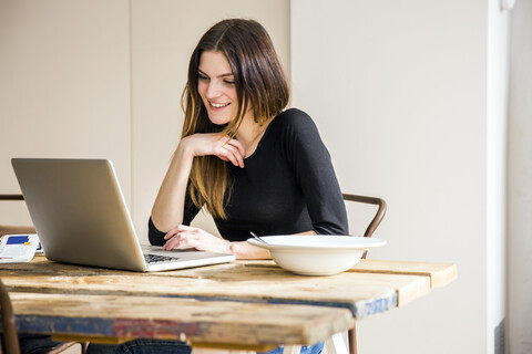 Junge Frau sitzt am Tisch in der Wohnung beim Frühstück und liest am Laptop, lizenzfreies Stockfoto