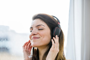Schöne junge Frau, die mit Kopfhörern vor einem Wohnungsfenster Musik hört - CUF16853