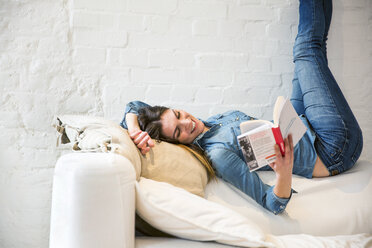 Junge Frau liegt mit hochgelegten Beinen auf dem Sofa und liest ein Buch - CUF16861