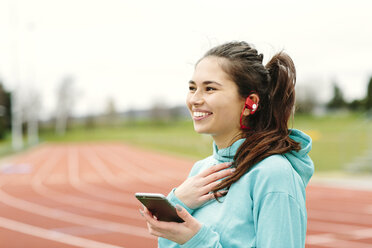 Young woman beside running track, holding smartphone, wearing earphones, smiling - CUF17182