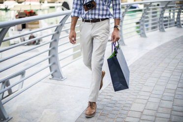 Waist down view of young man strolling on waterfront carrying shopping bag, Dubai, United Arab Emirates - CUF17223