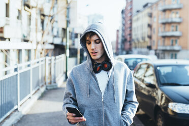 Man in urban area wearing hooded top and headphones looking down at smartphone - CUF17293