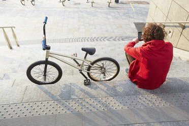 Rückansicht eines Mannes, der auf einer Treppe mit BMX sitzt und ein Smartphone benutzt - CUF17361