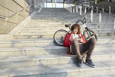 Mann sitzt auf Stufen mit BMX und benutzt Smartphone - CUF17363