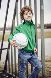 Boy leaning against railings holding football looking at camera smiling - CUF17382