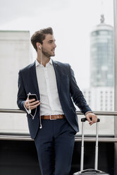 Young businessman at the window with cell phone, earbuds and rolling suitcase - DIGF04503