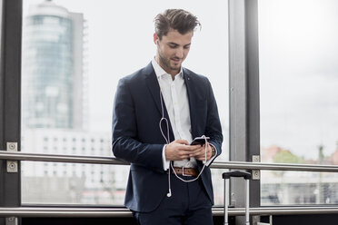 Young businessman at the window with cell phone, earbuds and rolling suitcase - DIGF04504