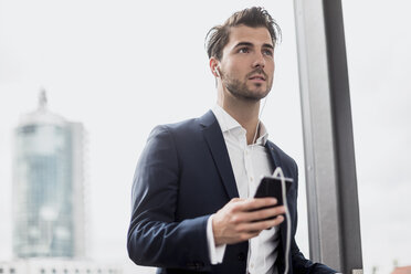 Businessman at the window with cell phone and earbuds - DIGF04505