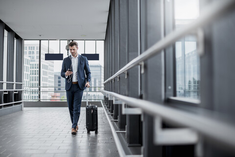 Young businessman in a passageway with cell phone, earbuds and rolling suitcase on the go stock photo