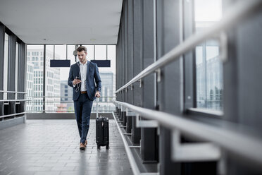Young businessman in a passageway with cell phone, earbuds and rolling suitcase on the go - DIGF04506