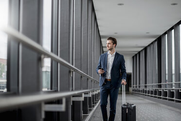 Young businessman in a passageway with cell phone, earbuds and rolling suitcase on the go - DIGF04510