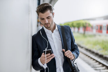 Geschäftsmann am Bahnhof mit Ohrstöpseln und Mobiltelefon - DIGF04522