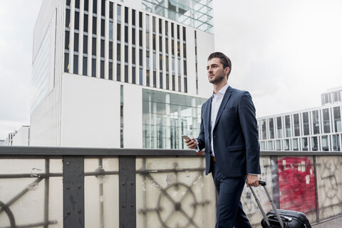 Young businessman with cell phone and rolling suitcase in the city on the go stock photo