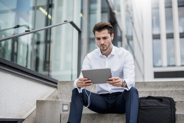 Businessman sitting on stairs with earbuds and tablet - DIGF04529