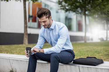 Young businessman sitting outdoors with takeaway coffee checking the time - DIGF04535