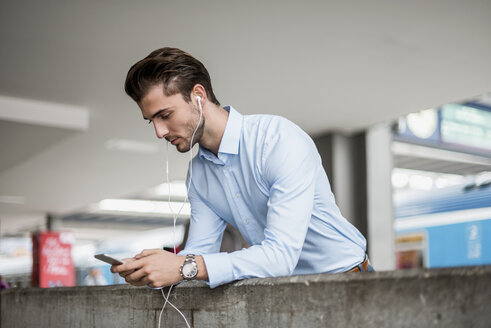 Geschäftsmann mit Mobiltelefon und Kopfhörern am Bahnhof - DIGF04553