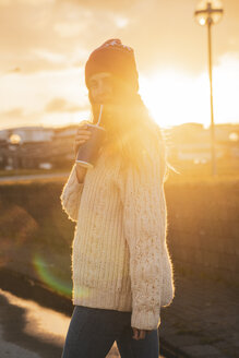 Island, junge Frau mit Kaffee zum Mitnehmen bei Sonnenuntergang - KKAF01116