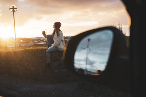 Iceland, young woman with coffee to go at sunset, wing mirror stock photo