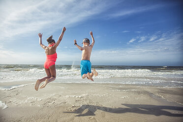 Rear view of girl and boy on beach arms raised jumping in mid air - ISF06468