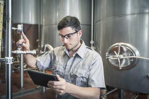 Junger Mann in einer Brauerei, der ein digitales Tablet in der Hand hält und die Anzeige überprüft, lizenzfreies Stockfoto