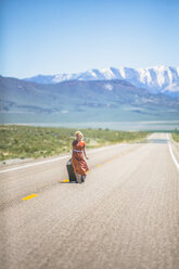 Junge Frau im Stil der 1950er Jahre, die allein auf dem Highway 50 spazieren geht und einen Koffer auf Rädern zieht, Nevada, USA - ISF06576