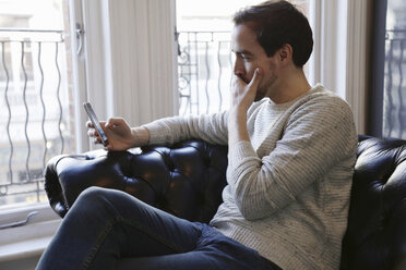 Mittlerer erwachsener Mann sitzt auf dem Sofa und schaut auf sein Smartphone - CUF17409