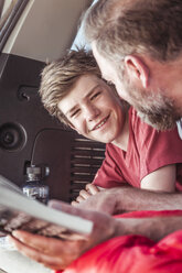 Mature man and teenage son reading guide book whilst lying in vehicle - CUF17413