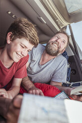 Mature man and teenage son reading map whilst lying in vehicle - CUF17415