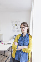 Young woman leaning against wall in office arms crossed looking at camera - CUF17750