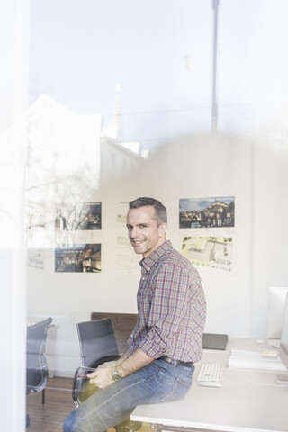 Blick durch Glas, Seitenansicht eines reifen Mannes, der am Schreibtisch sitzt und lächelnd in die Kamera schaut, lizenzfreies Stockfoto