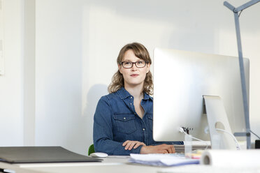 Junge Frau mit Brille sitzt am Schreibtisch und benutzt einen Computer, schaut weg und lächelt - CUF17788