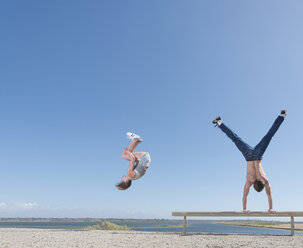 Girl doing somersault, boy doing handstand on beach bench - ISF06716