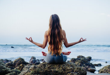 Rear view of young woman with long hair practicing lotus yoga pose on rocks at beach, Los Angeles, California, USA - ISF06788