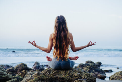 Rückansicht einer jungen Frau mit langen Haaren, die die Lotus-Yoga-Pose auf Felsen am Strand übt, Los Angeles, Kalifornien, USA - ISF06788