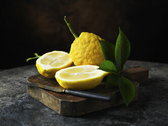 Strukturierte Zitrone, ganz und halbiert, mit Blättern auf einem Schneidebrett aus Holz mit einem alten Messer - CUF18016