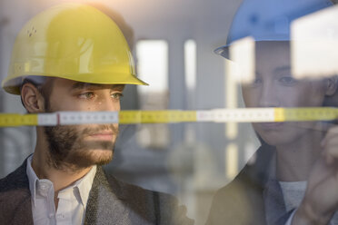 Young businessman and woman wearing hard hats measuring window in new office - CUF18112