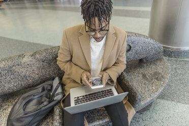 Junger Geschäftsmann sitzt im Bahnhof und benutzt Smartphone und Laptop - ISF06903