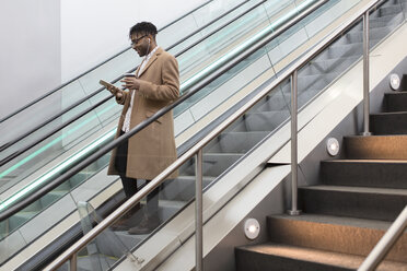 Junger Geschäftsmann, der die Rolltreppe eines Bahnhofs hinunterfährt und sein Smartphone liest - ISF06906