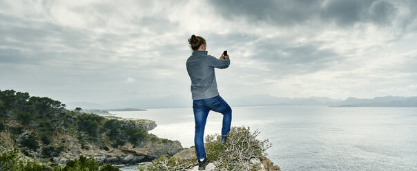 Panoramic view of young man standing on cliff photographing with smartphone, Alcudia, Majorca, Spain - CUF18418