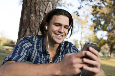 Young man sitting by tree wearing earphones looking at smartphone smiling - ISF06946