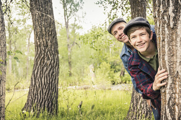 Junge und Mann im Wald mit flachen Mützen, die lächelnd hinter einem Baum hervorschauen - ISF06957