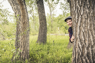 Jugendlicher im Wald mit flacher Mütze, der lächelnd hinter einem Baum hervorschaut - ISF06958