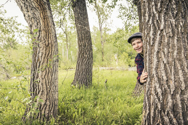 Teen boy in forest wearing flat caps peeking out from behind tree smiling - ISF06958